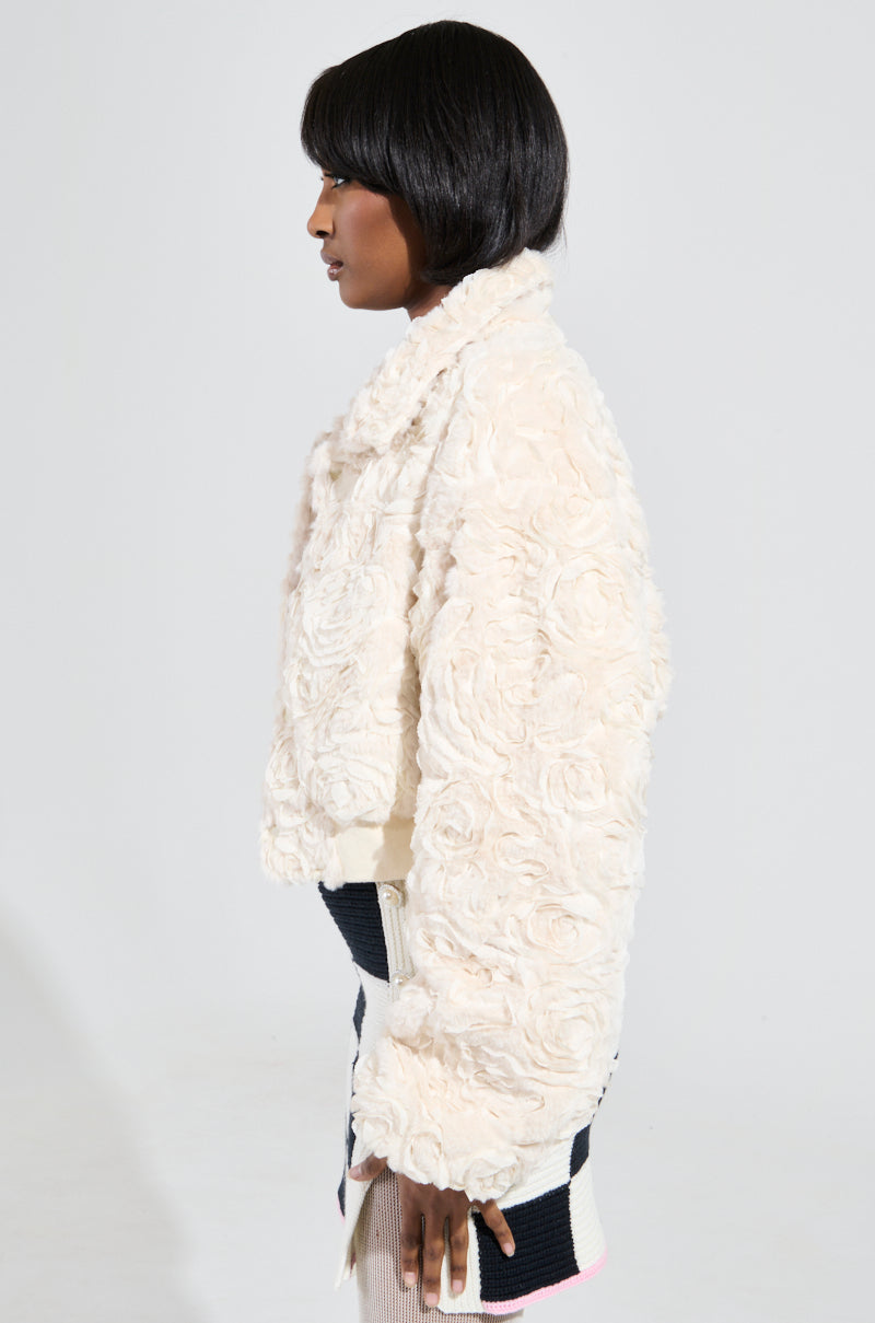 A person is standing in profile against a plain background, wearing a cream-colored FLORA TEXTURED BOMBER featuring a floral pattern paired with a patterned skirt. Their hair is styled in a sleek bob.