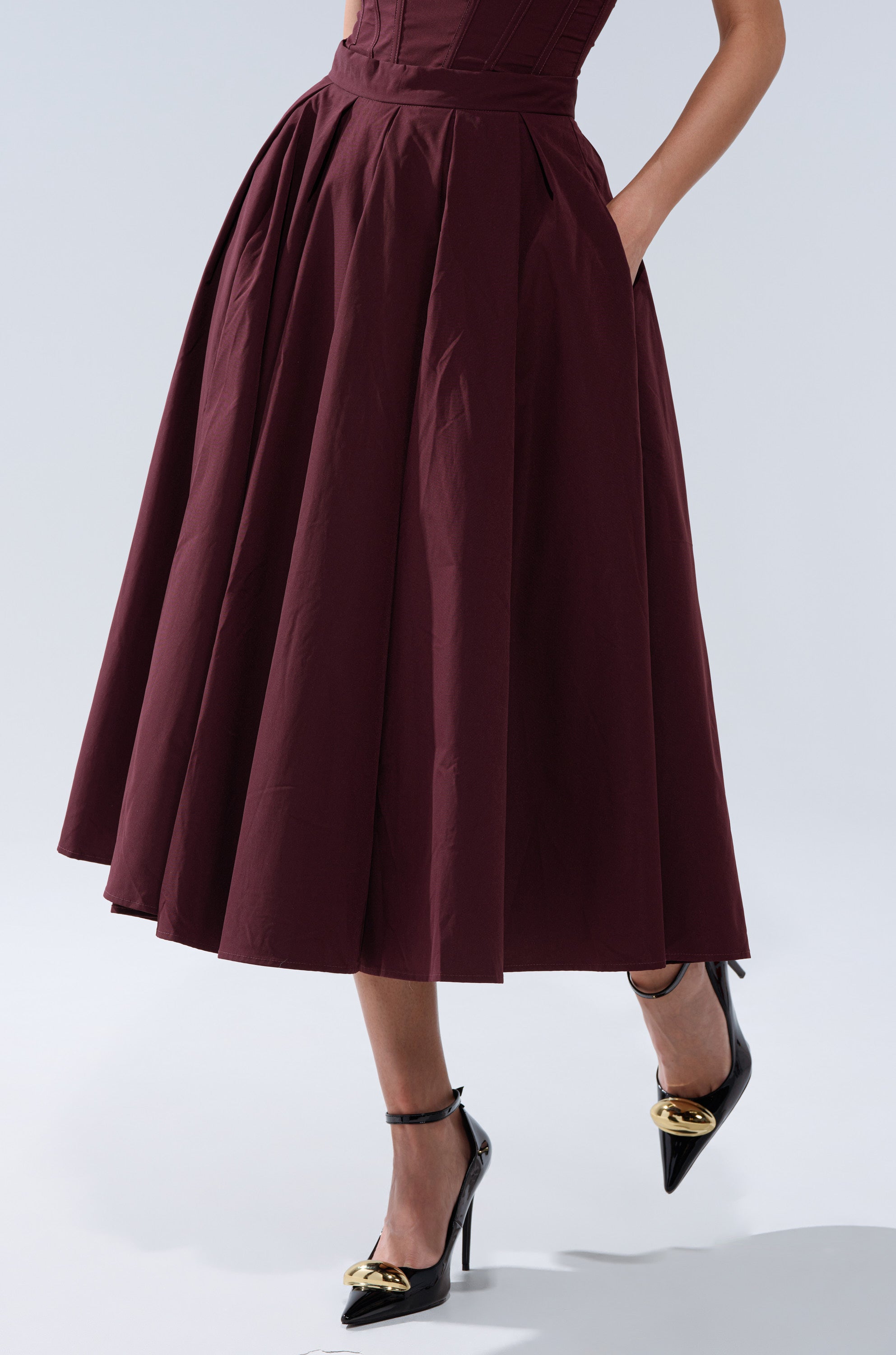 A person wearing the LOVE THIS FOR YOU MAXI SKIRT IN BROWN with their hand in a pocket and black pointed-toe heels with gold embellishments stands against a light background. 1