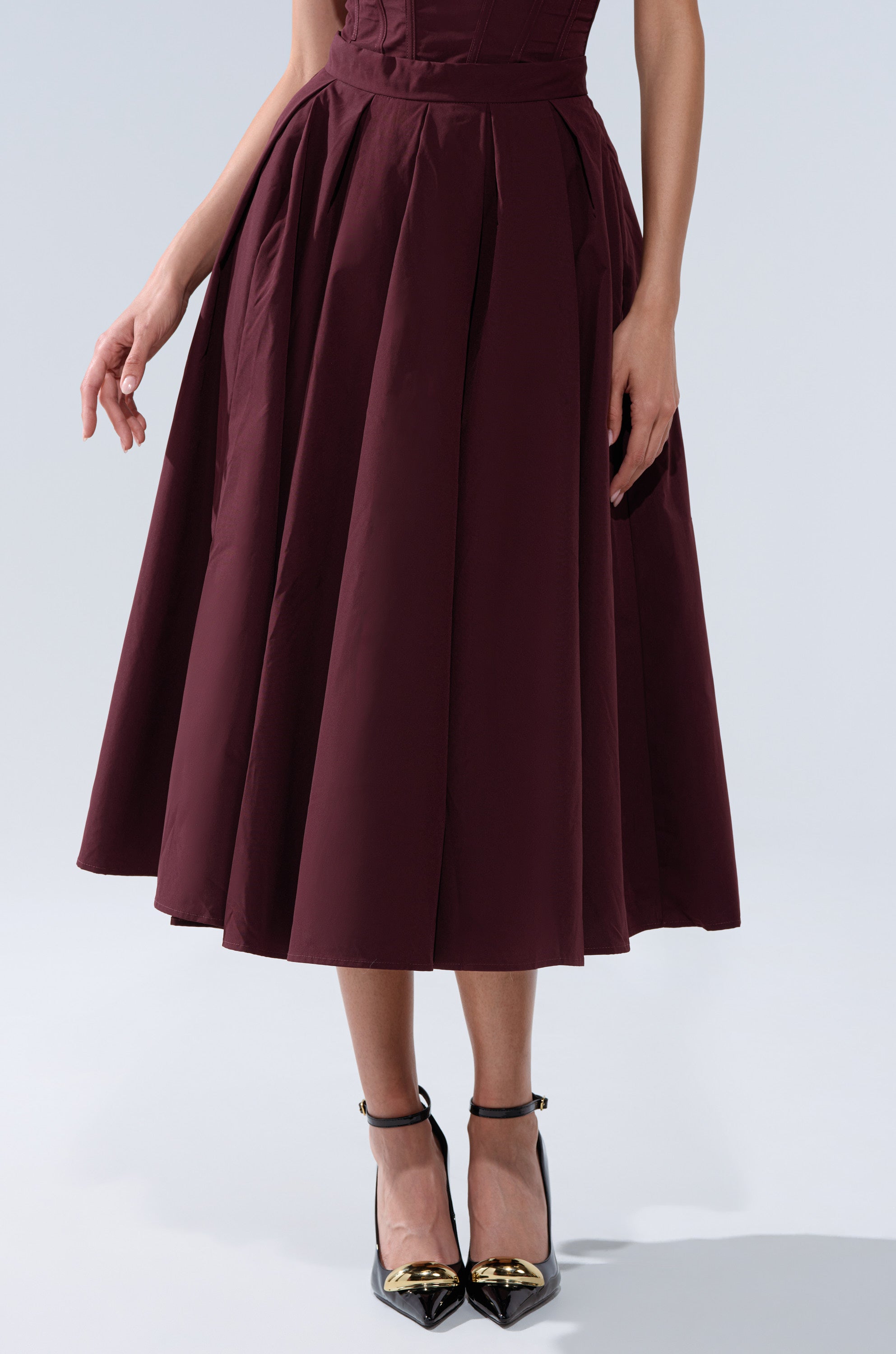 A woman wearing the LOVE THIS FOR YOU MAXI SKIRT IN BROWN, paired with black ankle-strap heels with gold pointed toes, stands against a light gray background. 2