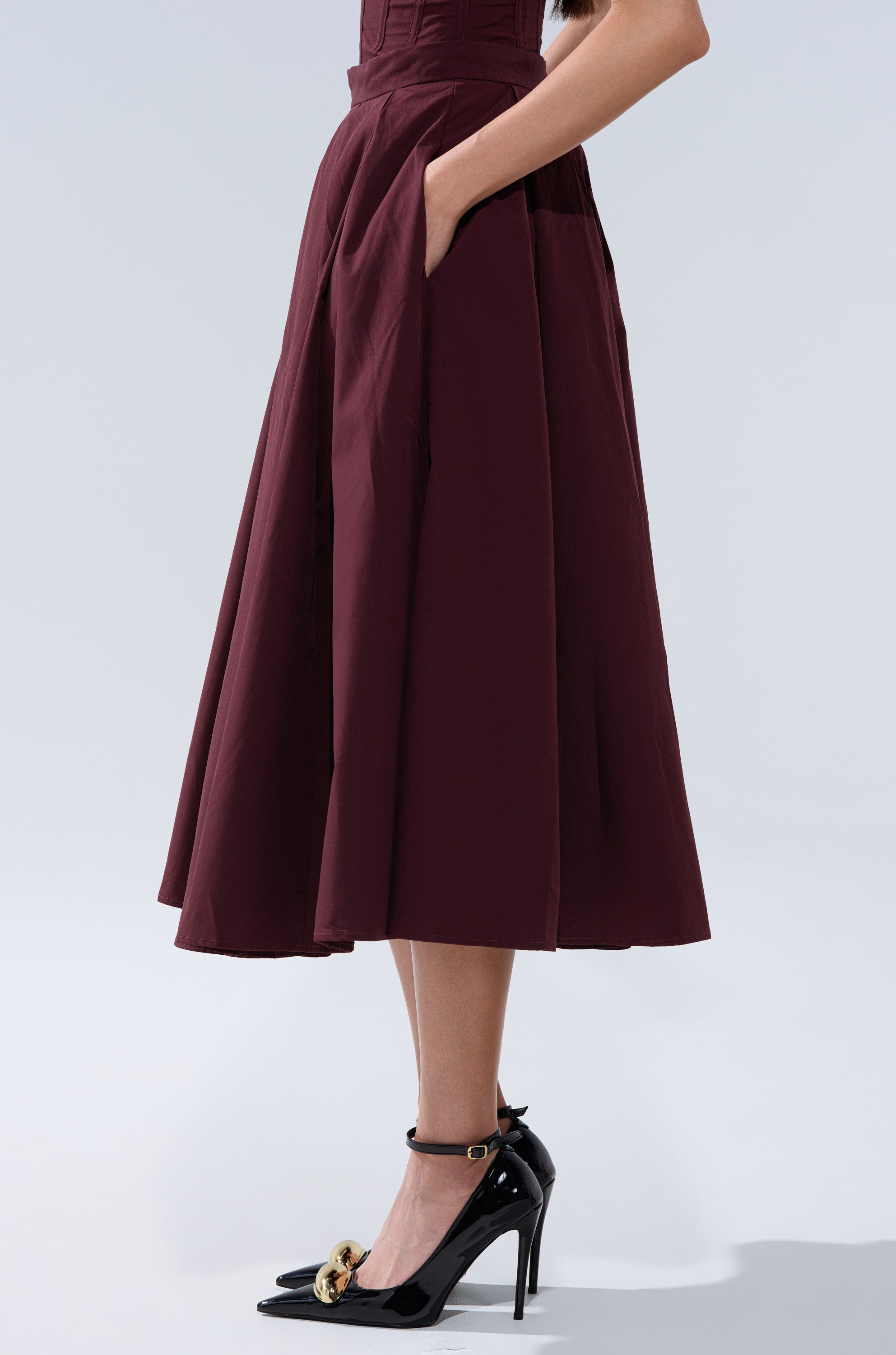 Someone wears the LOVE THIS FOR YOU MAXI SKIRT IN BROWN, a high-waisted, pleated midi skirt with pockets, styled with black patent leather heels featuring gold accents against a plain light background. 3