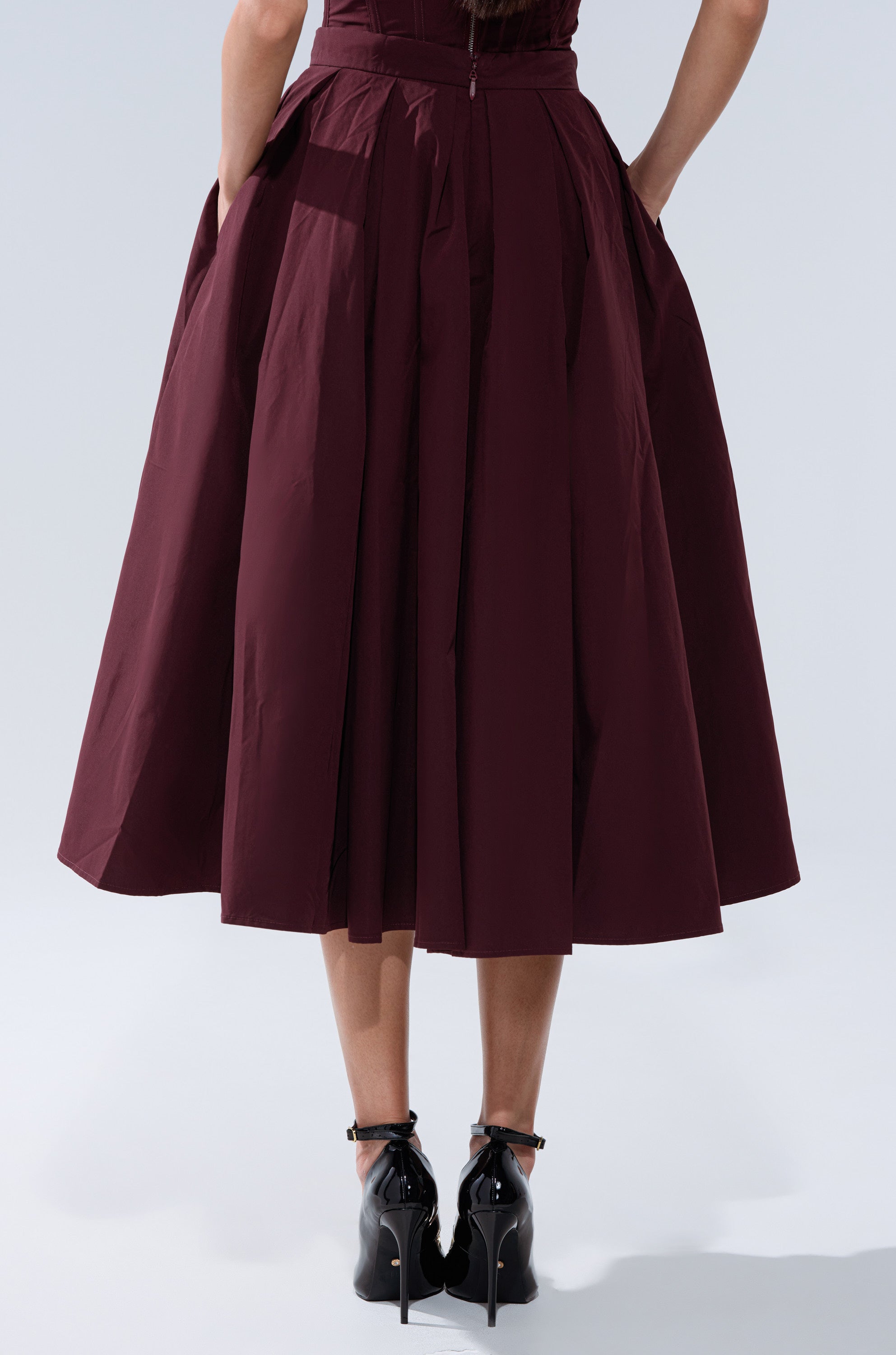 A woman stands with her hands in the pockets of the LOVE THIS FOR YOU MAXI SKIRT IN BROWN. She wears black, shiny high-heeled shoes with ankle straps. The photo is taken from behind. 4
