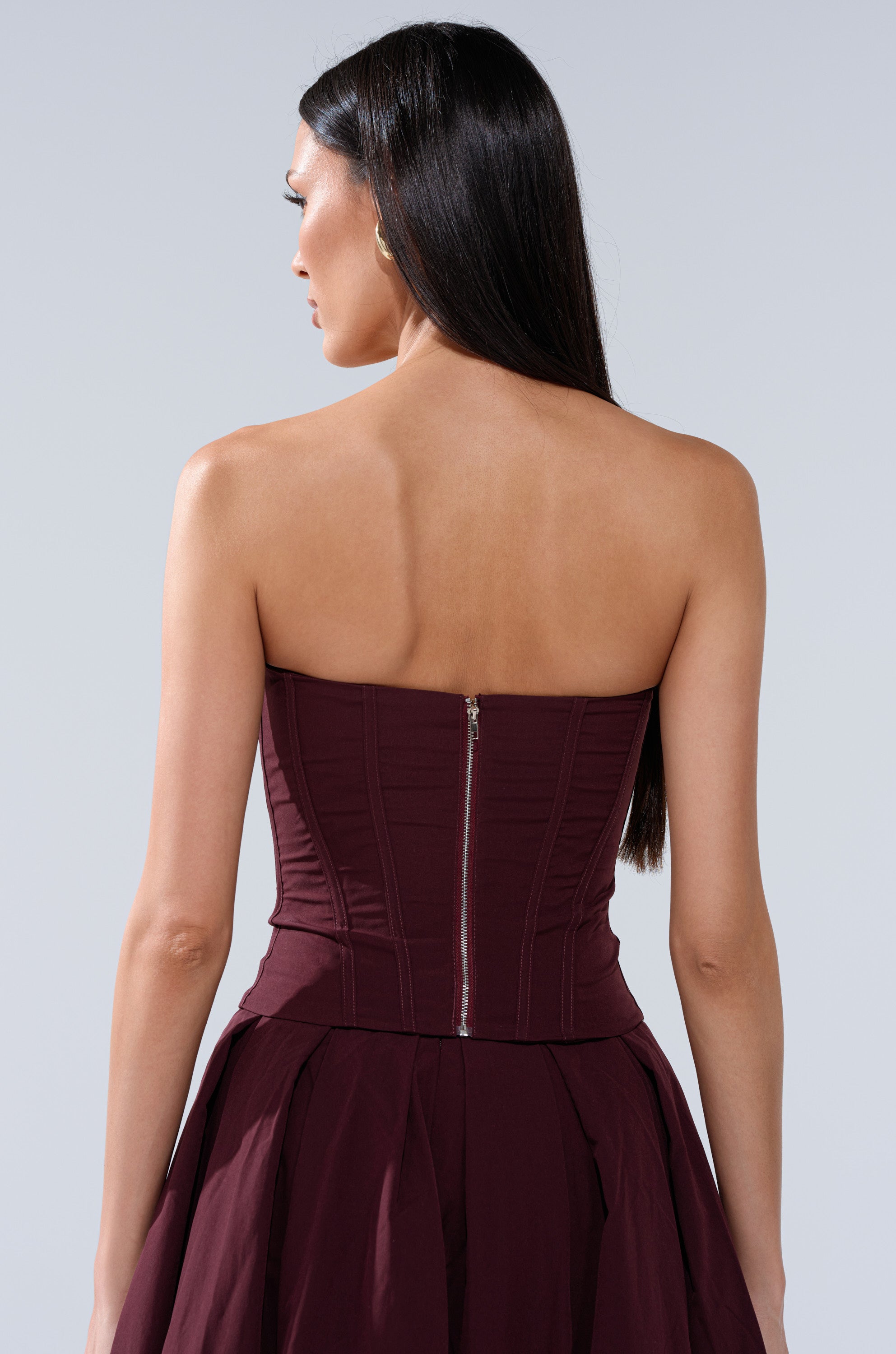 A woman with long dark hair is seen from the back in the LOVE THIS FOR YOU CORSET IN BROWN, a strapless dress with a sweetheart neckline, visible zipper on the fitted bodice, and structured boning, set against a plain light background. 3