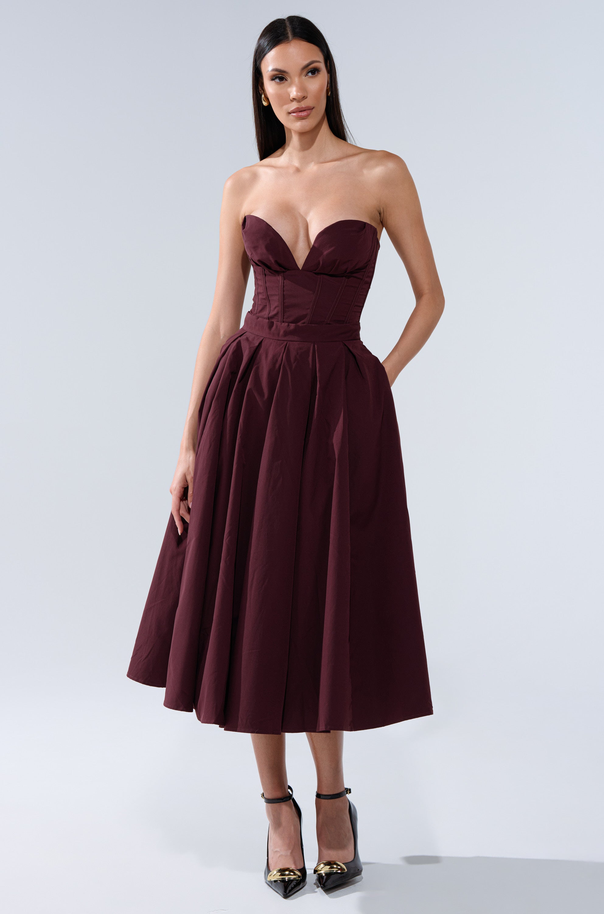 A woman stands against a plain light gray background wearing the LOVE THIS FOR YOU CORSET IN BROWN, styled as a strapless midi dress with a sweetheart neckline and structured boning, paired with black ankle-strap heels. 4