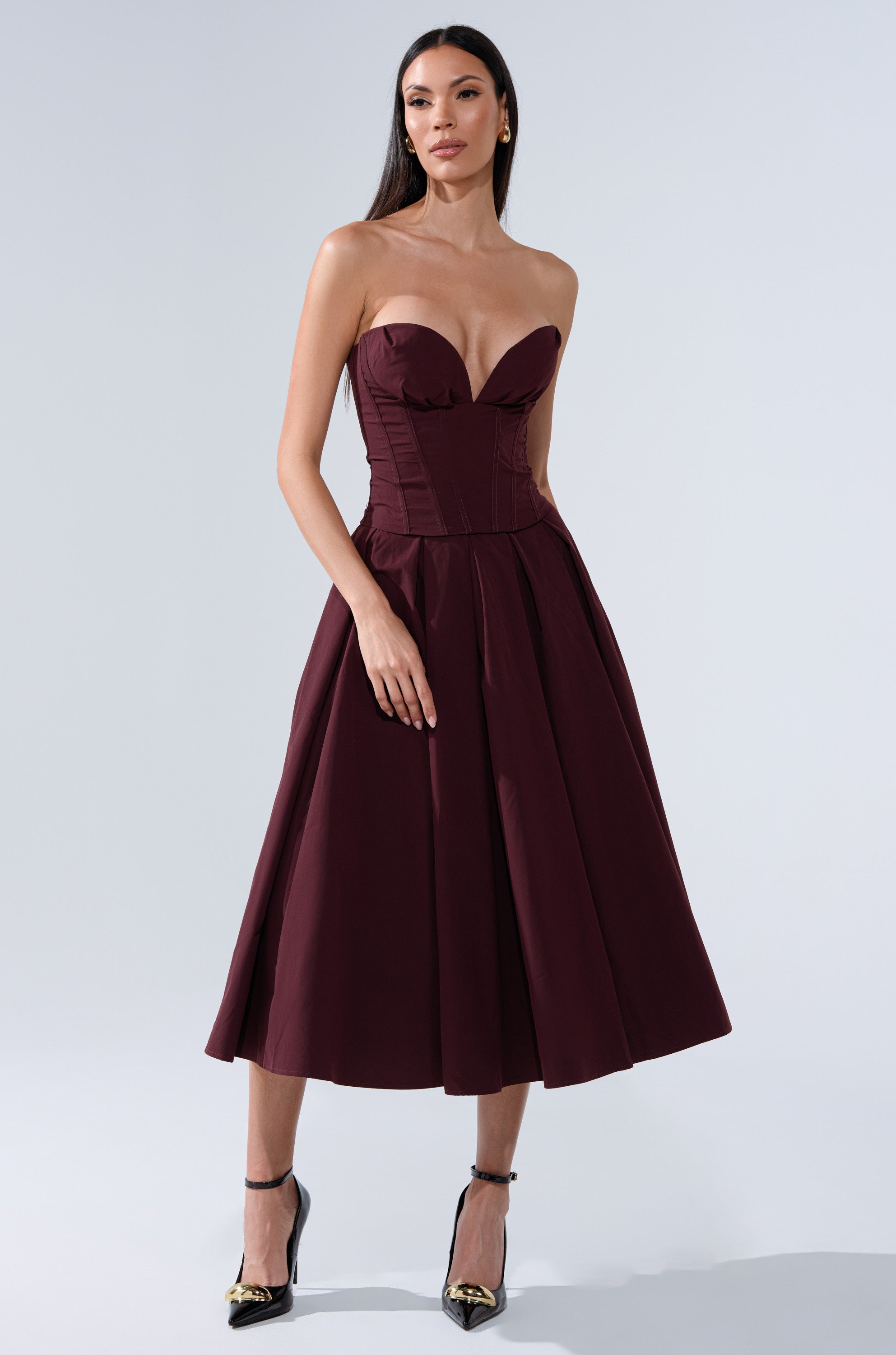 A woman in the LOVE THIS FOR YOU CORSET IN BROWN, a strapless midi dress with structured boning and a sweetheart neckline, stands against a plain light background, paired with black heels featuring gold tips. 5