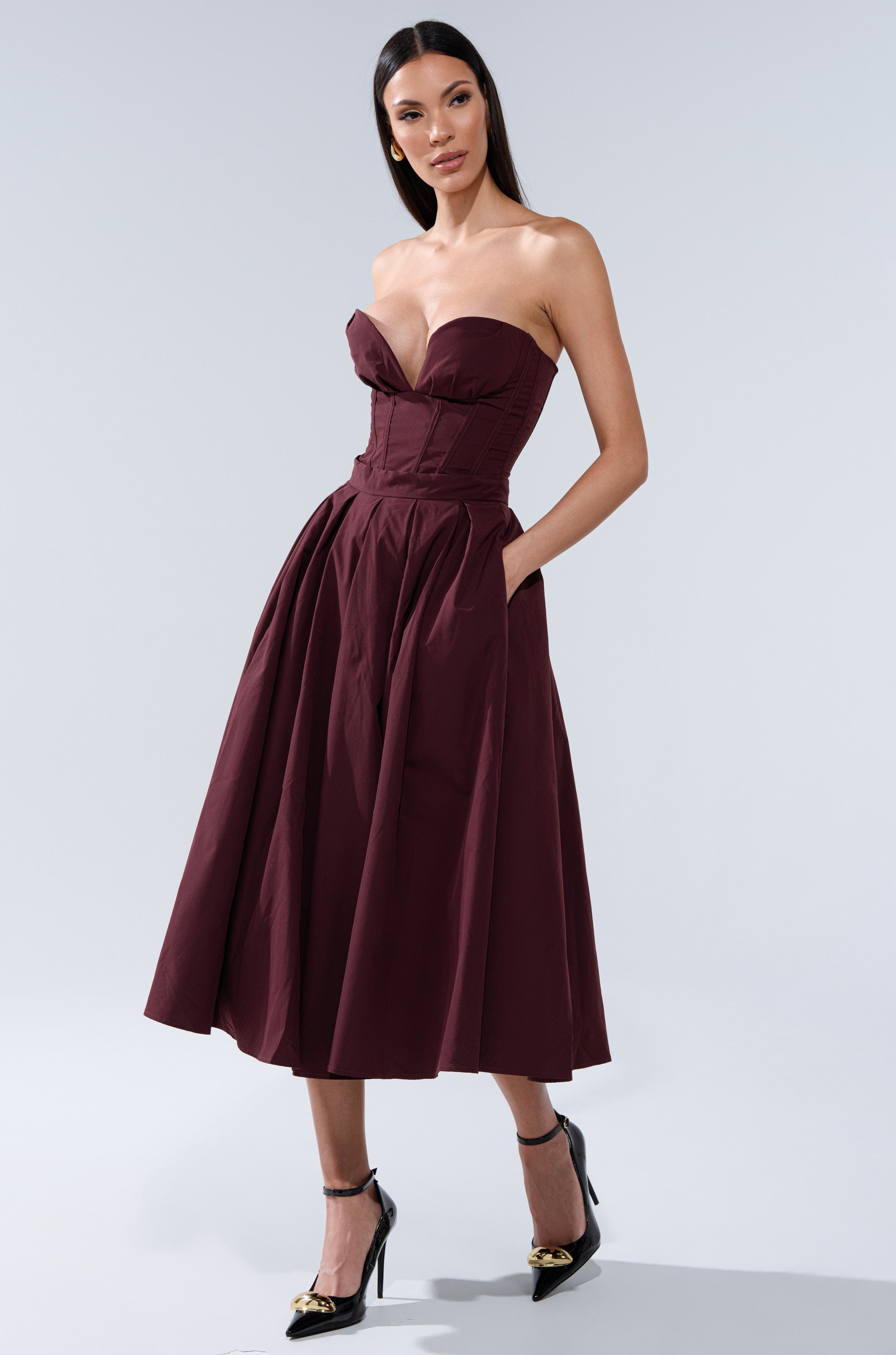 A woman models the LOVE THIS FOR YOU CORSET IN BROWN, a strapless, fitted piece with a sweetheart neckline and flared skirt. She pairs it with black pointed-toe heels featuring gold accents. Her long hair is straight against a plain light gray background. 7