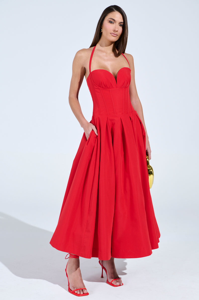 A woman poses confidently in a DOLCE DREAMING MIDI DRESS WITH CORSET. The strapless red dress features a flared skirt and sweetheart neckline. She holds a small gold purse and wears matching red high-heeled sandals against a plain white background.