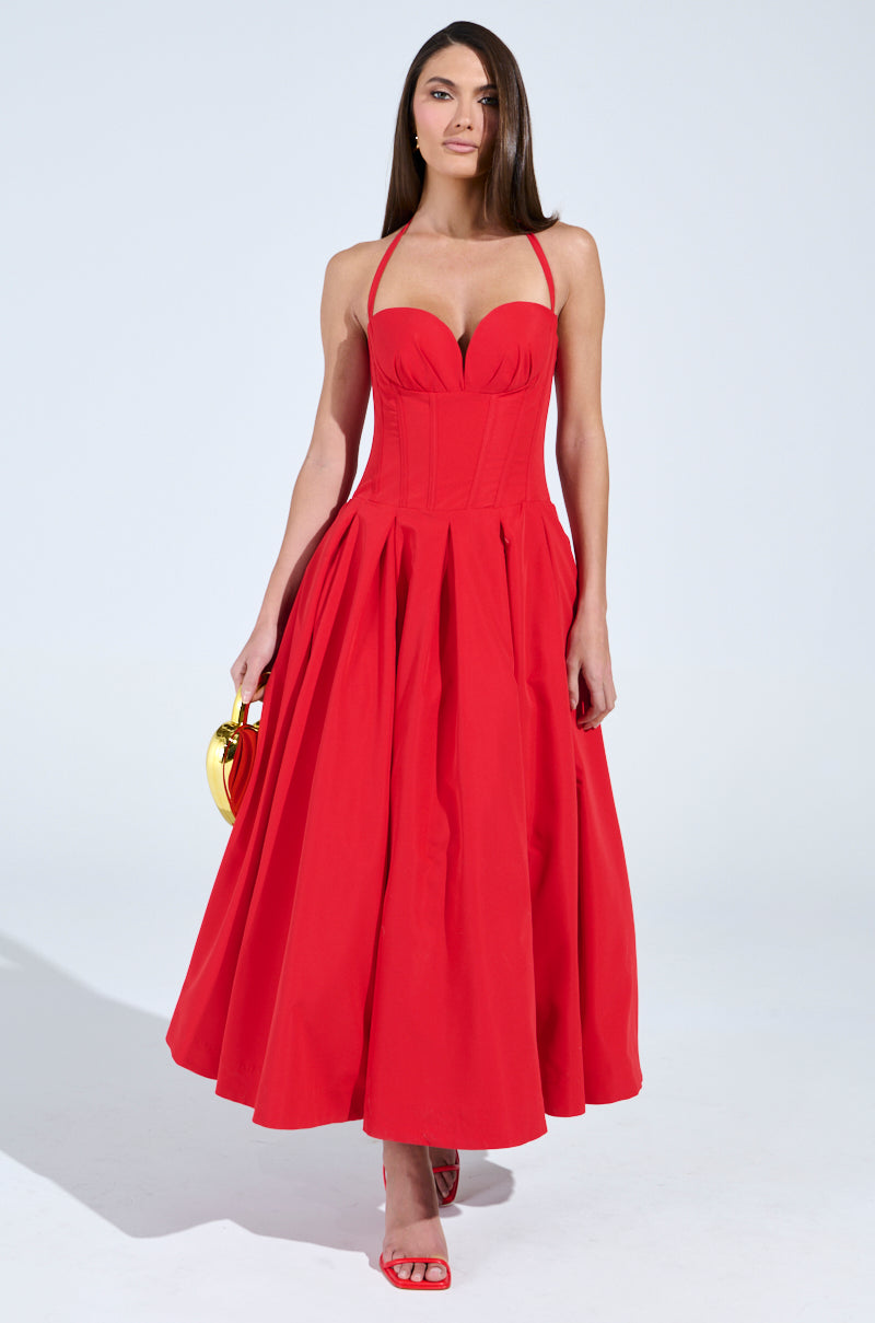 A woman poses confidently in a dazzling red DOLCE DREAMING MIDI DRESS WITH CORSET featuring a sweetheart neckline. She holds a small gold handbag and wears matching red high-heeled sandals. The plain white background highlights her striking ensemble.