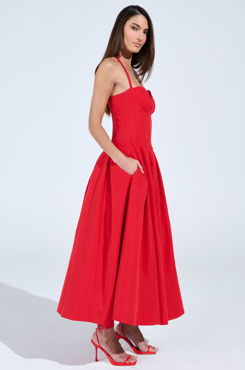 A woman stands confidently in the vibrant red DOLCE DREAMING MIDI DRESS WITH CORSET by AKIRA Label, featuring pockets and a sweetheart neckline. She pairs it with matching high heels, her elegance highlighted against a plain white background.