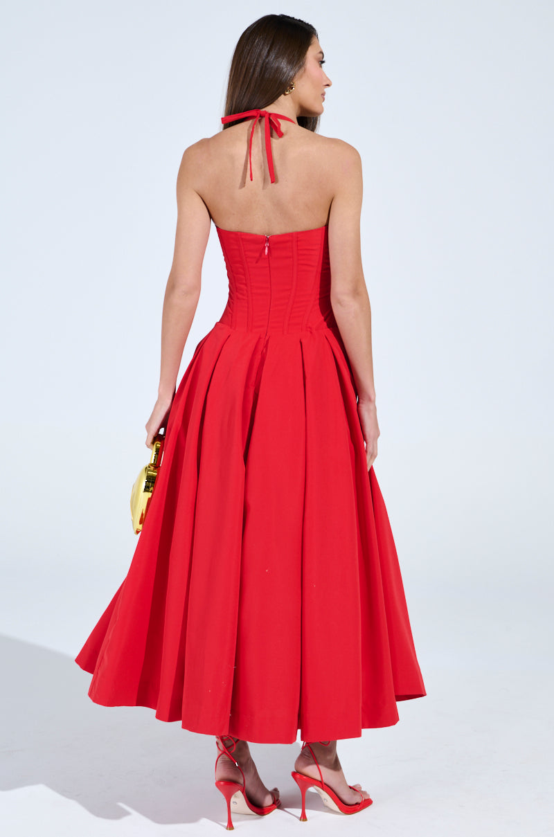 A woman, back to the camera, wears a sleeveless halter-neck red dress with a full skirt, resembling the DOLCE DREAMING MIDI DRESS WITH CORSET. She's holding a small gold handbag and sporting matching red high heels against a plain white background.