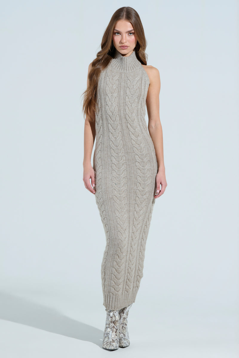 A woman with long brown hair stands against a plain backdrop in the JESSICA CABLE KNIT SWEATER MIDI DRESS, a sleeveless gray design featuring a high neck and chunky cable patterns that reach her ankles, paired with snake-patterned ankle boots.