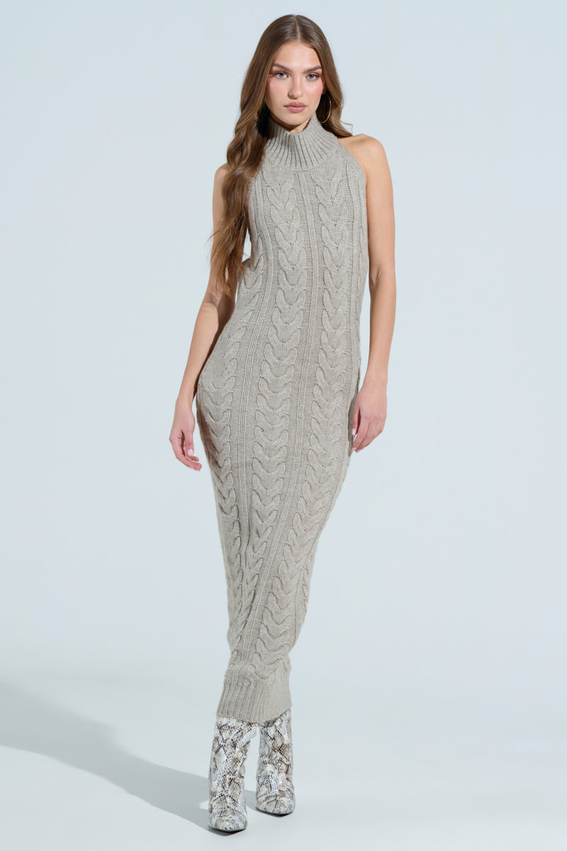 A person with long hair is wearing the JESSICA CABLE KNIT SWEATER MIDI DRESS, featuring a sleeveless, high-neck design and ankle-length chunky cable knit pattern. They pair it with snakeskin-patterned heeled boots against a plain light blue background.