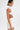 A person with short dark hair wears the NIGHT ON THE TOWN STRAPPY MINI DRESS—textured, white, strappy, with side cutouts. They have a bold red-pink abstract shoulder tattoo and hold a white clutch against a plain white background.
