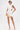 A woman in the NIGHT ON THE TOWN STRAPPY MINI DRESS—sleeveless, textured white with side cutouts—pairs clear high heels and gold earrings, holding a transparent clutch with a gold accent. Tattoos show on her left arm and thigh.