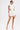 A woman models the NIGHT ON THE TOWN STRAPPY MINI DRESS—sleeveless, white, with textured fabric and side cutouts. She pairs it with a clear handbag, clear heels, and flaunts her skull tattoo for a chic night-out look.