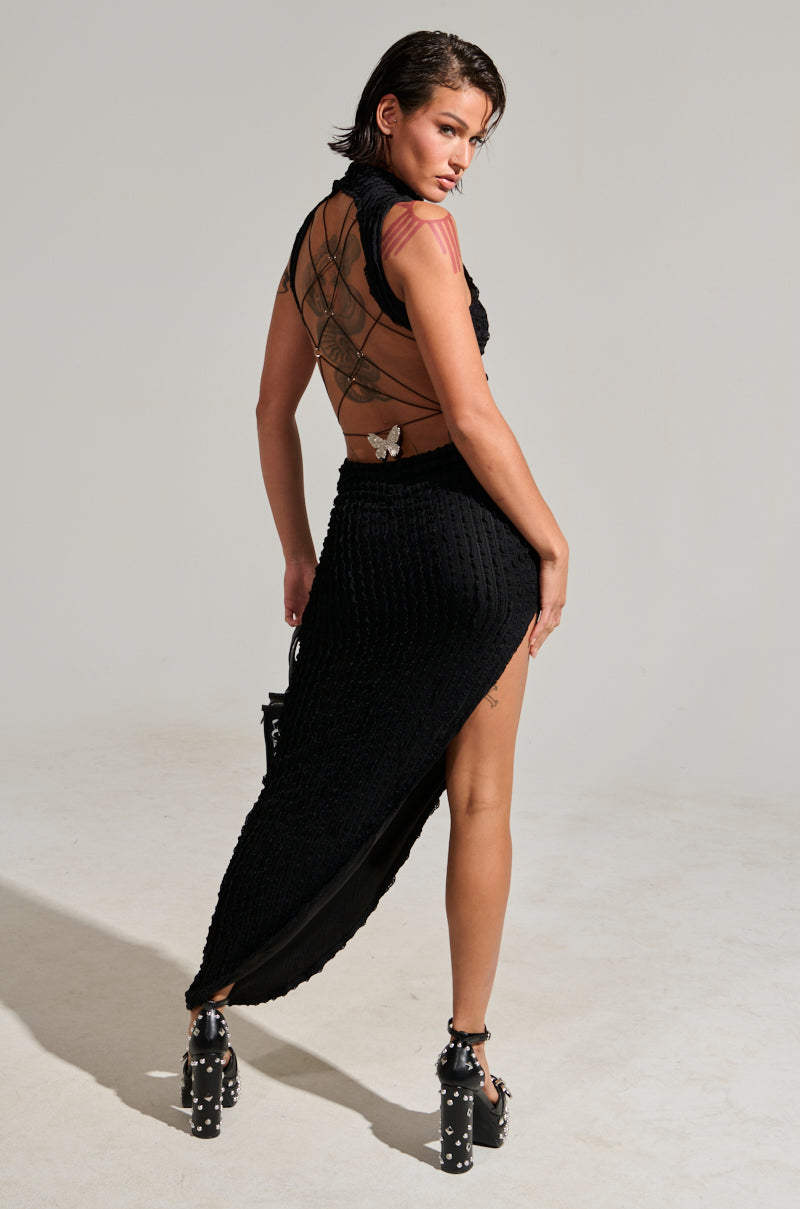 In a well-lit studio, a woman with short hair poses in the LET LOOSE MESH MAXI DRESS, showcasing its textured black fabric and high-slit mesh design with a sheer back. The dress is adorned with butterfly details at the waist. She complements her look with studded black heels and a metallic silver pendant.