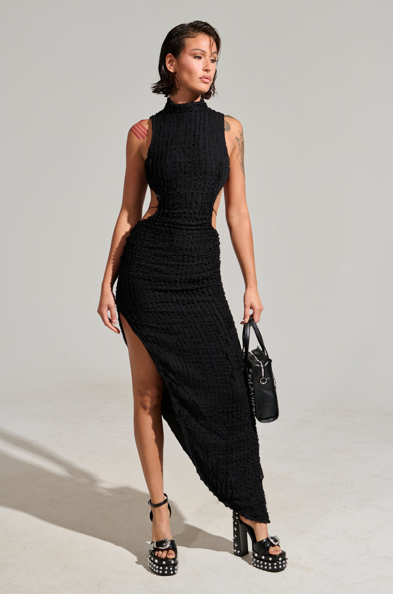 A person stands confidently in the LET LOOSE MESH MAXI DRESS, featuring a black textured fabric with an asymmetrical design and a high side slit. They accentuate the outfit with a metallic silver pendant, carry a sleek black handbag, and wear stylish platform heels. Tattoos are visible on their arms as they pose against the plain, neutral background.