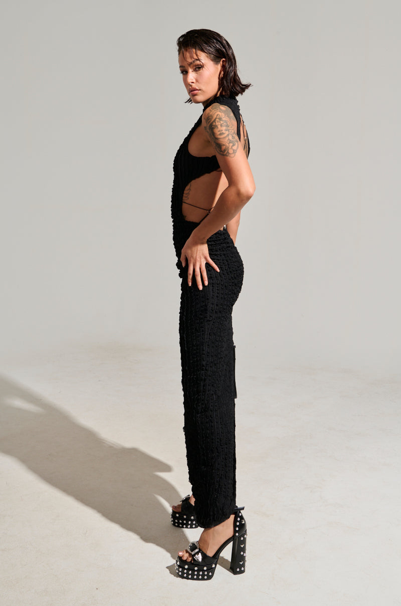 A person with short dark hair stands confidently in a side pose, wearing the LET LOOSE MESH MAXI DRESS in black, showcasing its sleeveless cut-out details, paired with platform shoes featuring metal decorations. A metallic silver pendant adds a touch of elegance against the plain, neutral background, highlighting their figure and attire.