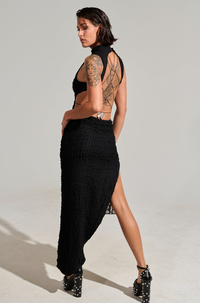 A person with short dark hair stands facing away, highlighting the LET LOOSE MESH MAXI DRESS in black. This textured, backless bodycon dress features a high slit. A large tattoo adorns their left arm and is paired with a metallic silver pendant. The ensemble is completed with black high-heeled shoes adorned with studs, set against a plain light gray background.