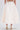 A person wears the long, ivory GIRL POWER MIDI SKIRT with an A-line silhouette and pearl-embellished high heels, standing against a plain light background. The cropped image shows only the lower half of the body from behind.
