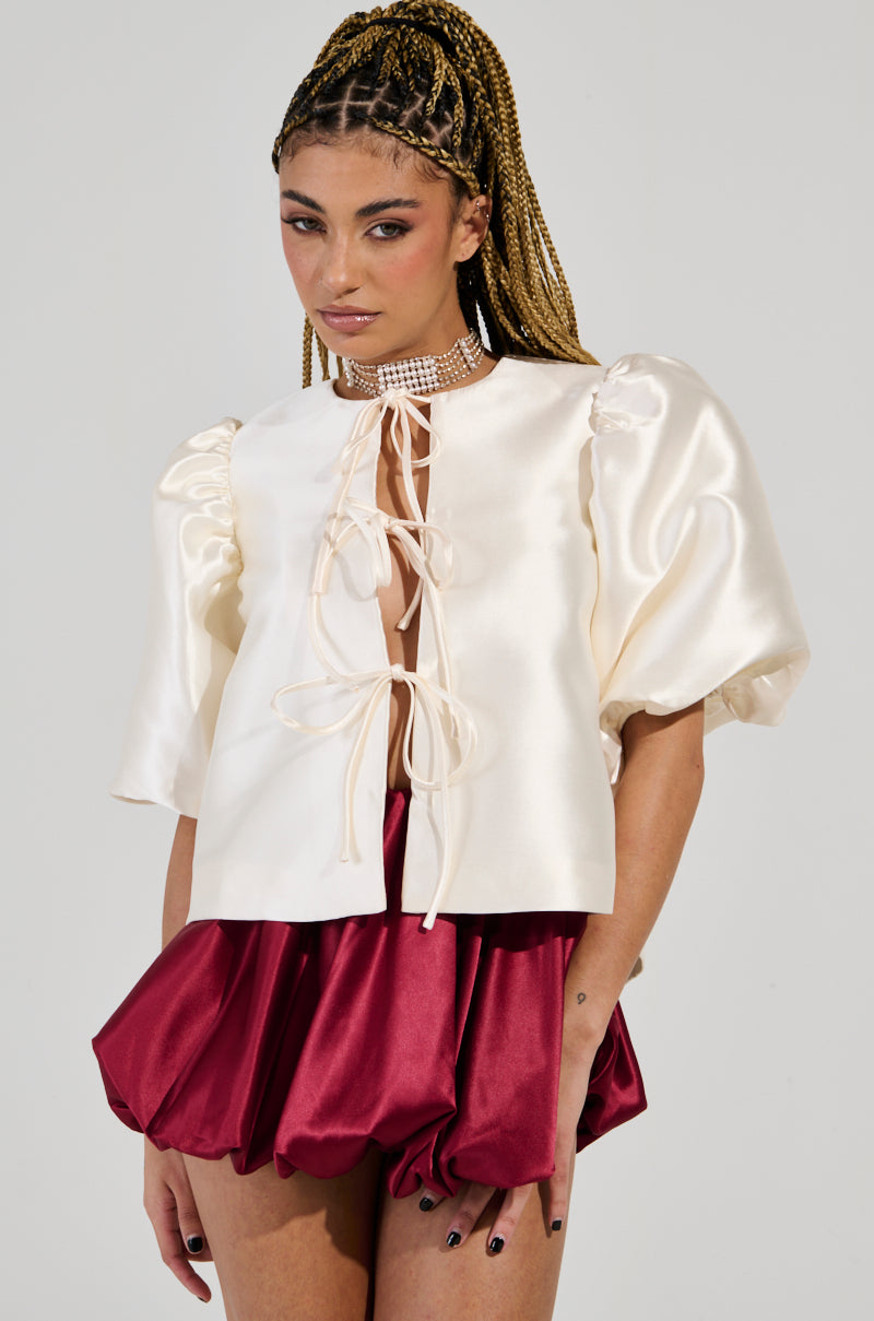 The model is wearing the ALL THE TEA TIE FRONT BLOUSE IN IVORY, which features puffed sleeves and a chic self-tie front closure. It's perfectly complemented by a vibrant red bubble skirt. She enhances the look with a dazzling choker and showcases her braided hair, all set against a simple white backdrop.