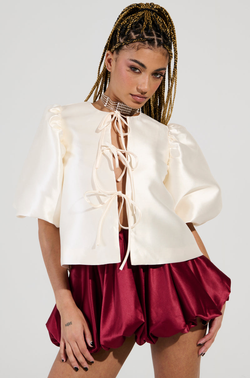 Wearing the chic "ALL THE TEA TIE FRONT BLOUSE IN IVORY," a person exudes confidence with its self-tie front closure and puffed sleeves. Complemented by burgundy, balloon-style shorts, their braided hair and a hand tattoo accentuate the ensemble against a plain gray backdrop.
