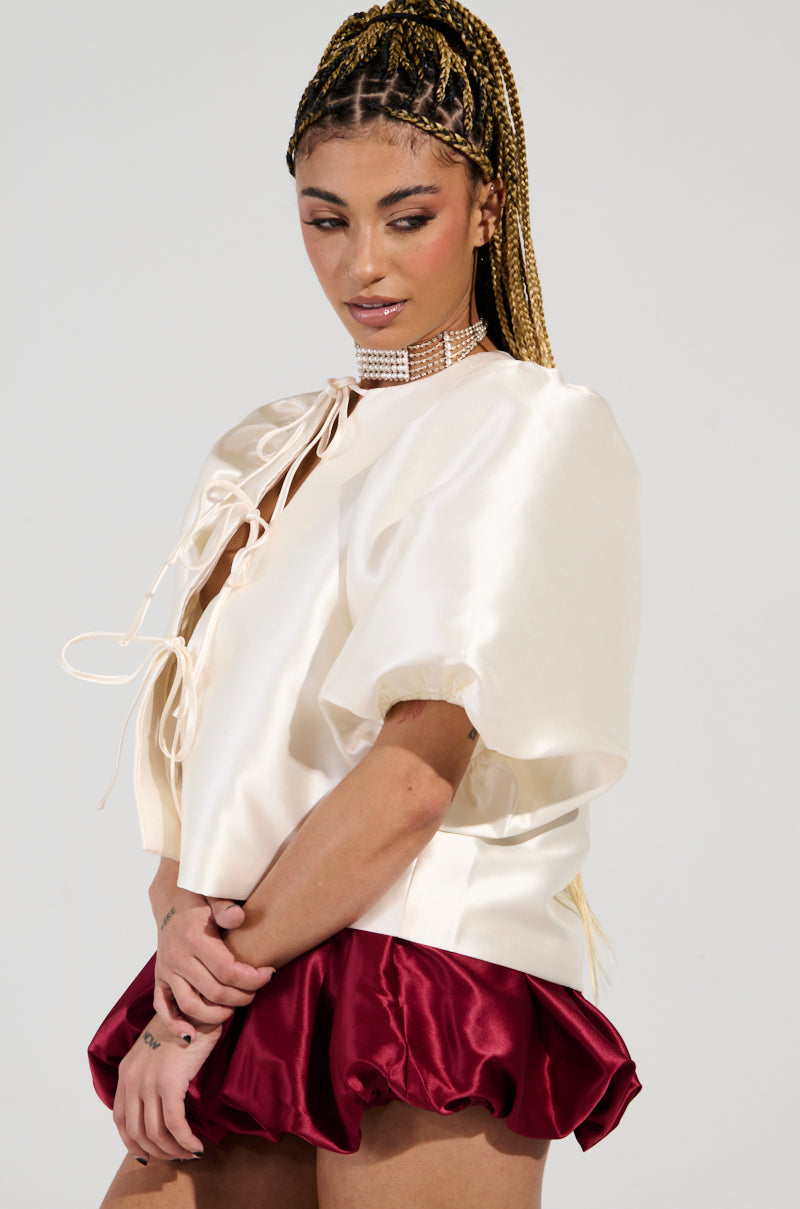 A person with braided hair is wearing the ALL THE TEA TIE FRONT BLOUSE IN IVORY, characterized by its puffed sleeves and self-tie front closure, paired with a maroon satin skirt. They accessorize with a pearl choker necklace while standing against a plain background.