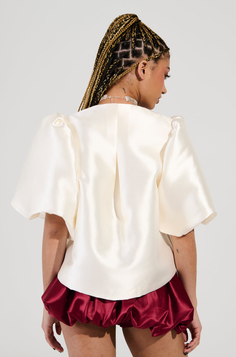 A person with braided hair is wearing the sophisticated "ALL THE TEA TIE FRONT BLOUSE IN IVORY," featuring puffed sleeves and a self-tie front closure, paired with a burgundy skirt. They are standing against a plain background, facing away from the camera.
