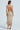 A woman poses in the GET YOU A ME WOVEN MAXI DRESS, featuring a textured, backless design with ruched detailing, thick straps, and a long fitted skirt. She pairs it with high-heeled sandals and rests her hand on her hip against a plain light background.
