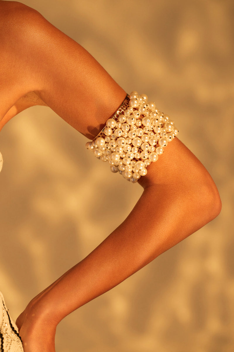 A tanned arm showcases the elegant PURRRL PLEASE BRACELET with clustered pearls against a softly lit backdrop, exuding warm sophistication.
