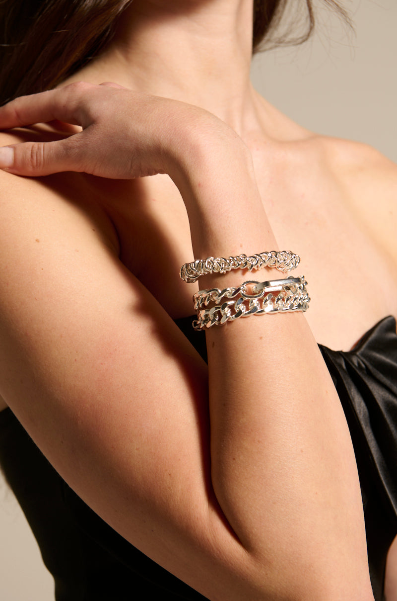 A person with long, dark hair wears a strapless black top. Their arm is adorned with a striking geometric-link bracelet from the IZZY SILVER BRACELET SET, featuring chunky and linked designs, resting against their shoulder in a posed manner.