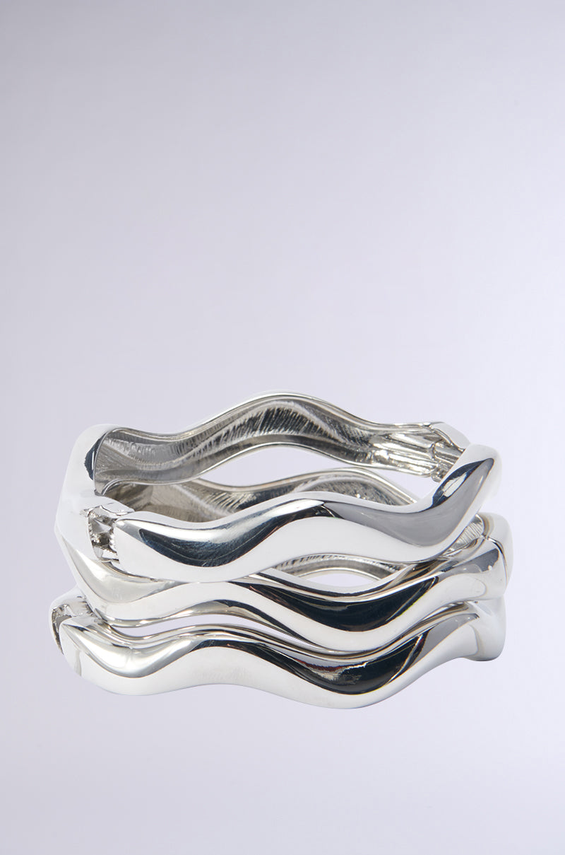 The HERE FOR THE VIBES BRACELET SET features three metallic faux silver bracelets with organic, wavy hammered silhouettes stacked against a plain background. Their reflective surface highlights a fluid and modern aesthetic, resulting in an elegant set that effortlessly catches the eye.