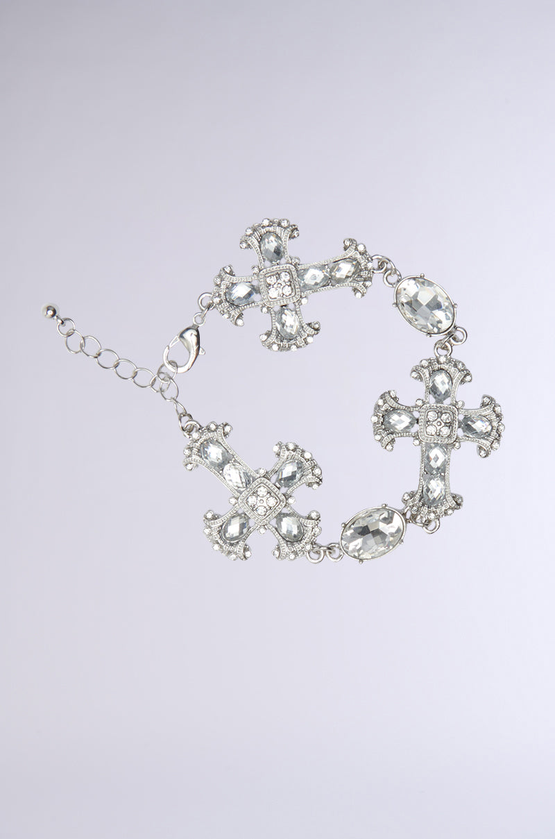 The "CROSS ME BRACELET" showcases detailed cross designs and oval gemstones, enhanced with shimmering rhinestone accents, set on a light gray base.