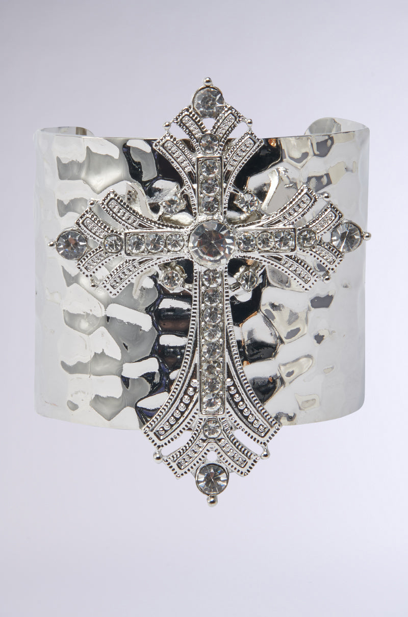 The GIVING ENERGY CUFF is a wide, hammered silver bracelet from the AKIRA Label with a central ornate cross pendant adorned with sparkling rhinestones, shown against a plain light background. 4