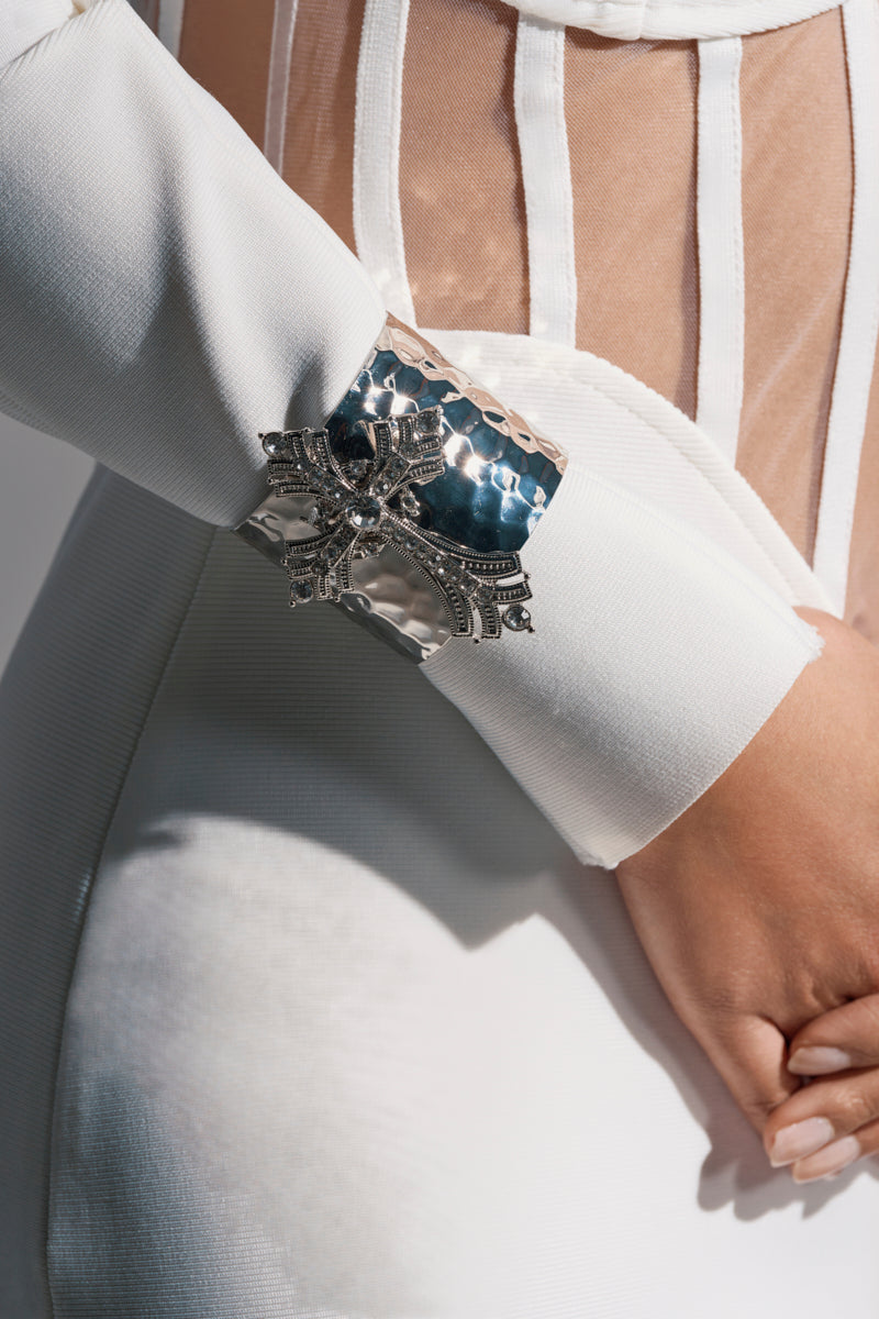 A close-up of a person’s arm in a white, long-sleeve AKIRA Label outfit with mesh panels, wearing the GIVING ENERGY CUFF—a large, ornate silver bracelet with textured metal and rhinestone embellishments—their hand resting on their hip.