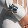 A close-up of a person’s arm in a white, long-sleeve AKIRA Label outfit with mesh panels, wearing the GIVING ENERGY CUFF—a large, ornate silver bracelet with textured metal and rhinestone embellishments—their hand resting on their hip.