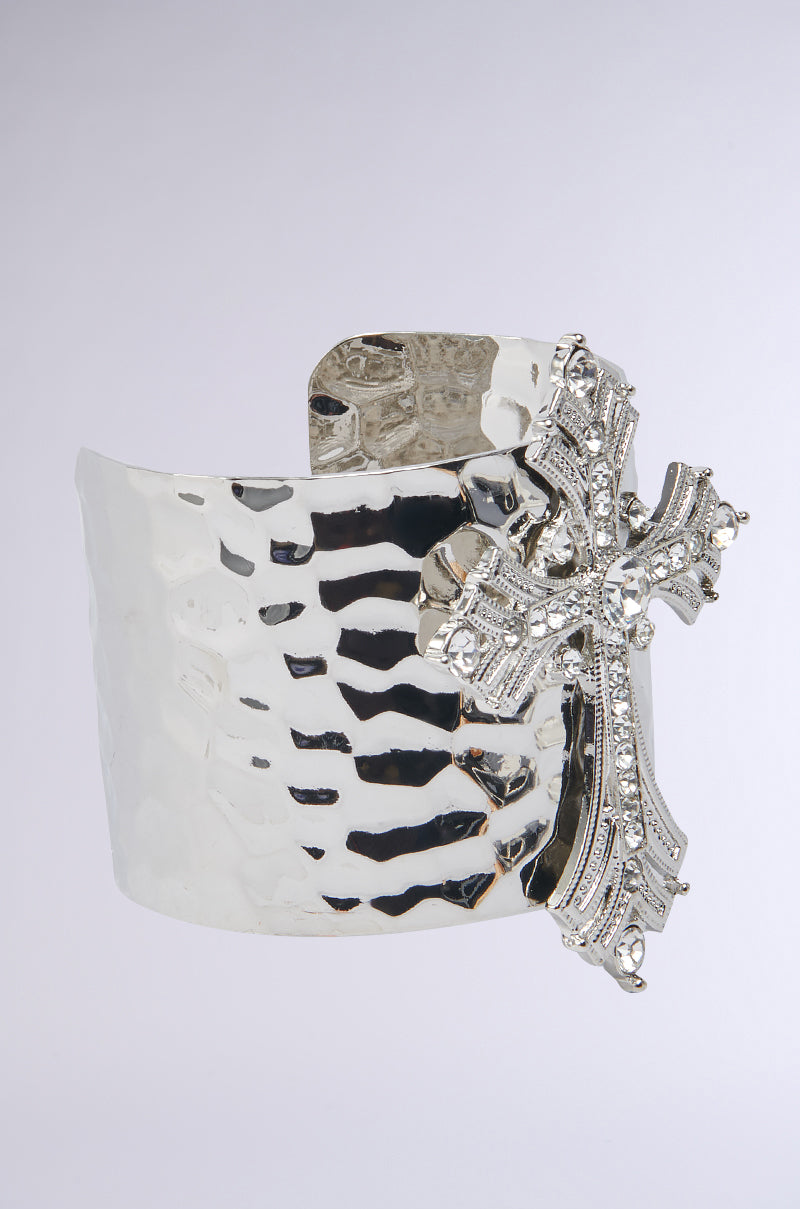 The GIVING ENERGY CUFF is a wide hammered silver bracelet from AKIRA Label, featuring a large ornate cross pendant embellished with sparkling rhinestones on the front.