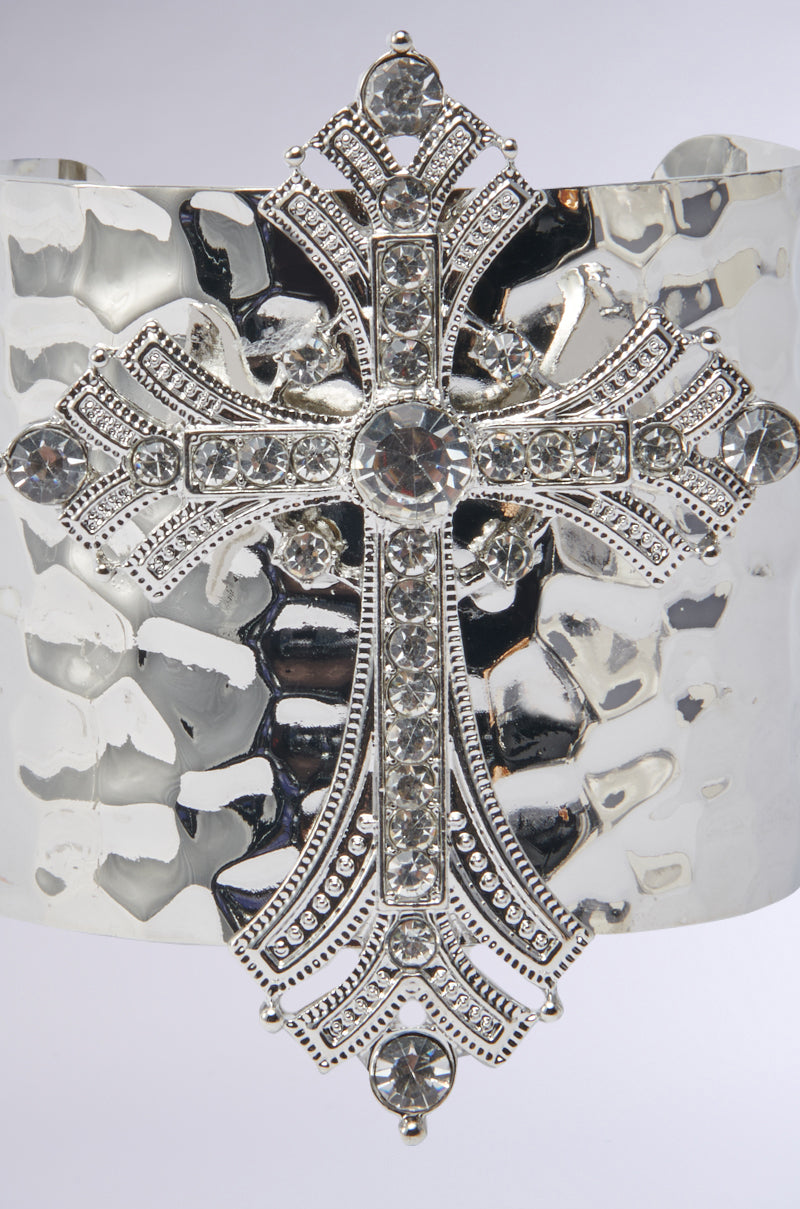 The GIVING ENERGY CUFF from AKIRA Label is a wide, hammered silver bracelet featuring an ornate cross pendant adorned with sparkling rhinestones at the center and on each arm.