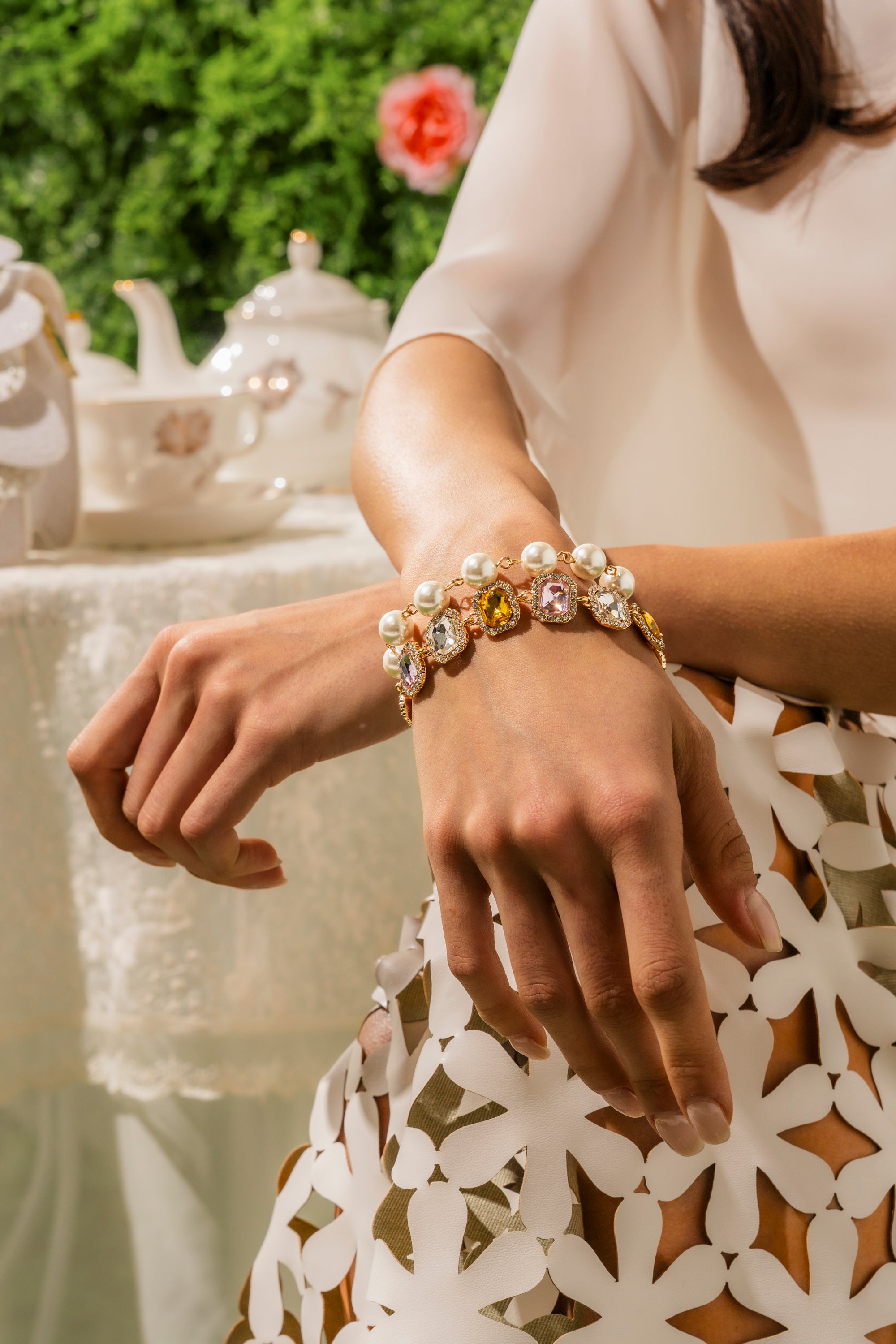A woman wearing the REGENCY BRACELET SET, featuring pearls and colorful gemstones on gold-tone links, sits in a white floral-cutout outfit at a tea table surrounded by greenery and flowers. 0