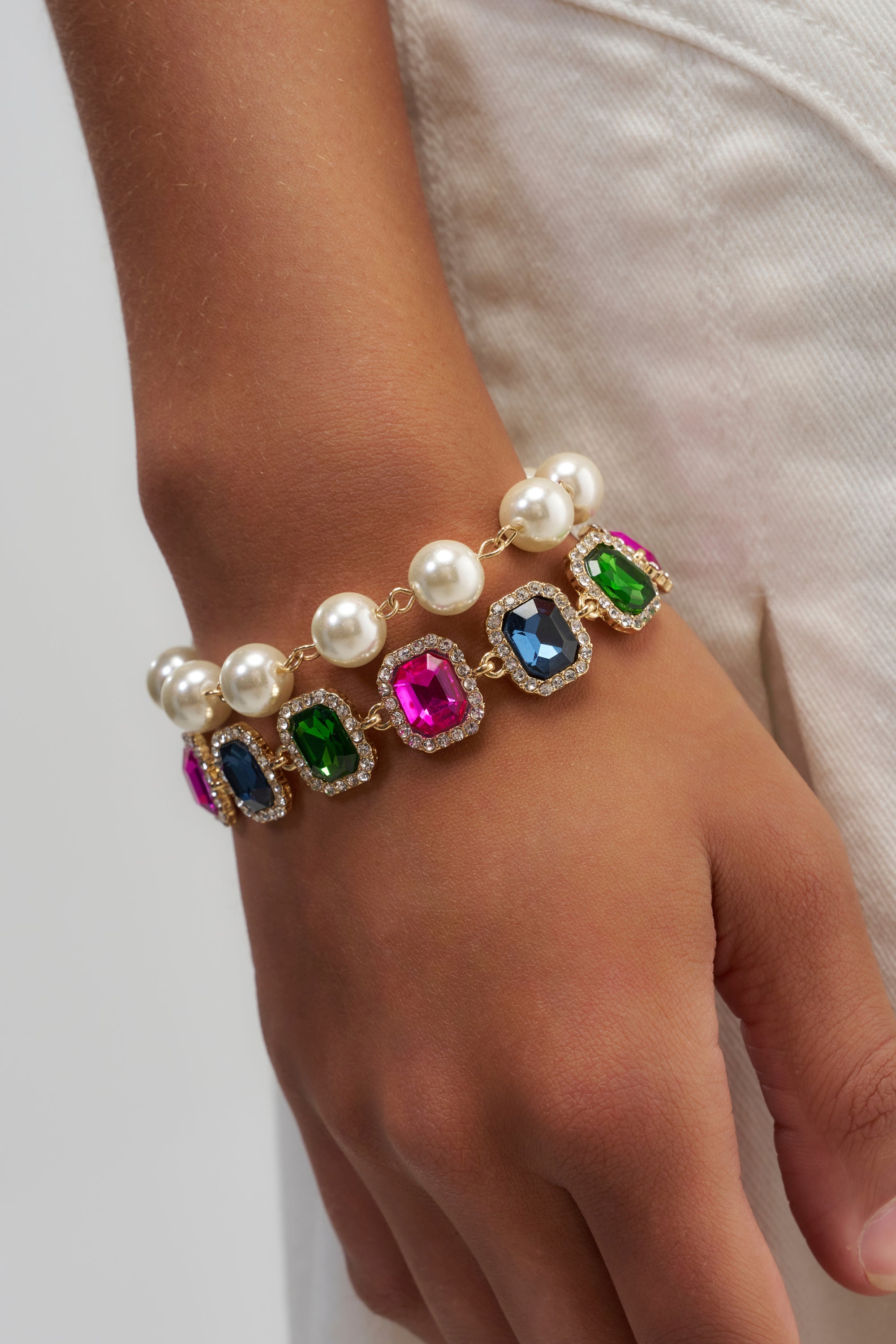 A hand adorned with the COLOR ME RICH BRACELET SET, featuring large faux pearls and colorful gemstones in shades of blue, green, and pink, rests beside white pants.