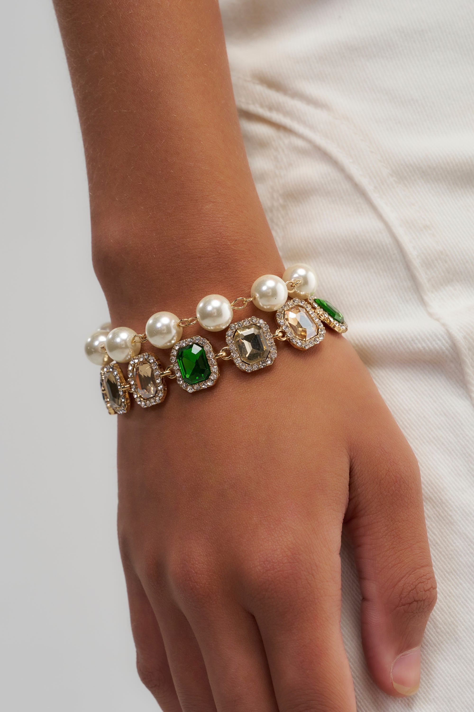 A hand wearing the QUEEN GREEN BRACELET SET, adorned with large faux pearls and rectangular green and clear gemstones, resting against white pants.