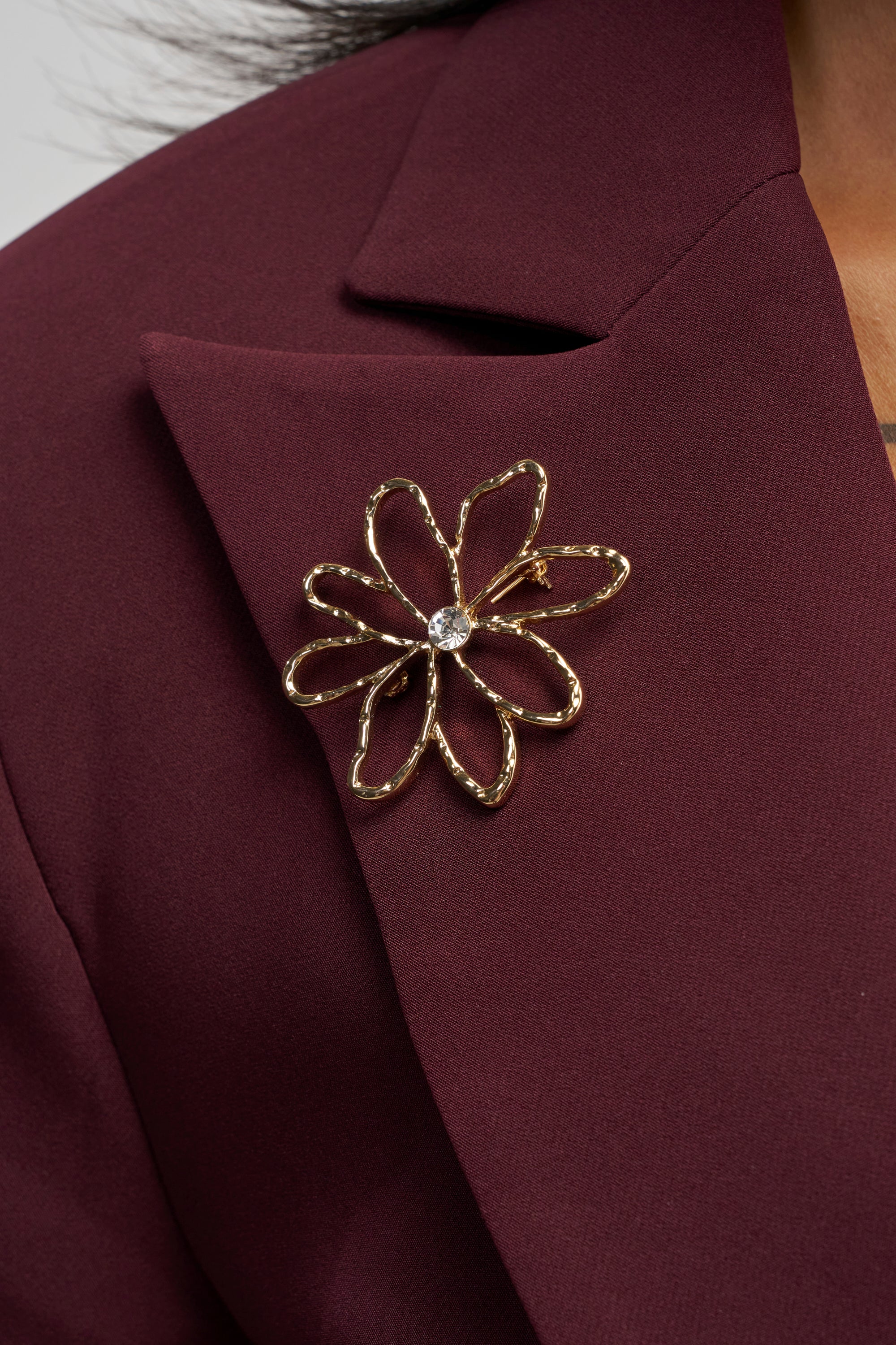 A FLORAL METAL BROOCH featuring a hammered gold flower design and a sparkling crystal rhinestone center is pinned to the lapel of a dark burgundy blazer.
