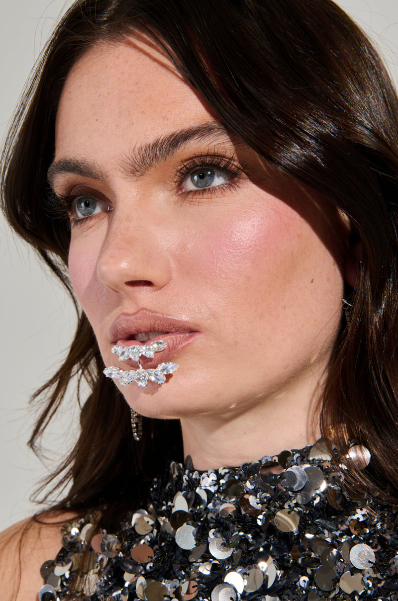 A woman with long, dark hair and shimmering gemstones on her lips sparkles in a high-neck sequined top. She accessorizes with the CHARMER FAUX LIP RING from AKIRA, which enhances the elegance of her thoughtful expression. The photograph portrays her in a well-lit setting. 1