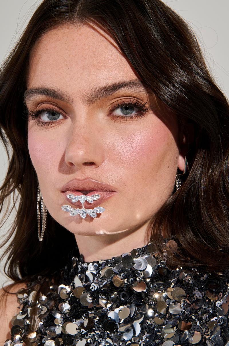 A woman with long brown hair is adorned in a sequined top and earrings. Her bold makeup features silver sequins shaped like a butterfly beneath her lips, enhanced by the CHARMER FAUX LIP RING. The neutral background makes her striking makeup and outfit stand out even more. 2