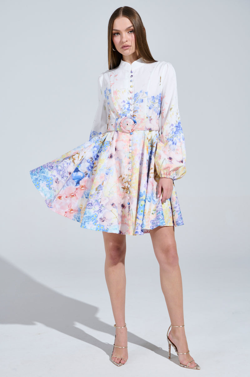 A woman confidently poses in a studio, wearing the EXPENSIVE TASTE FLORAL MINI DRESS—emphasizing her long straight hair and the colorful floral skater skirt. She gently lifts the skirt with one hand and completes her elegant look with strappy high heels.