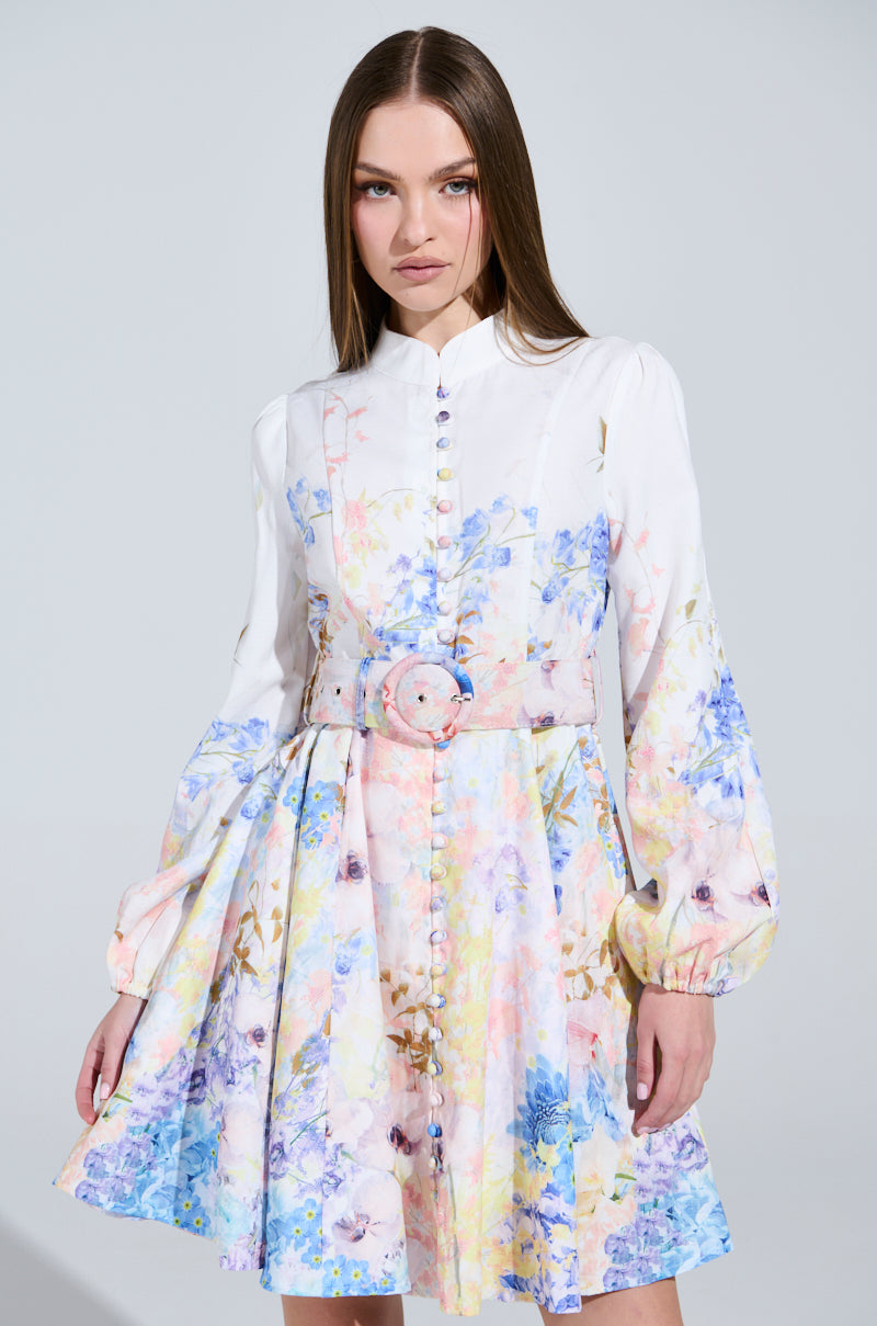 A person wearing the EXPENSIVE TASTE FLORAL MINI DRESS, featuring long sleeves, a button-up design, and a skater silhouette. The cinched waist with a matching belt gives an elegant flair to the mini dress. The scene is set against a plain gray background.