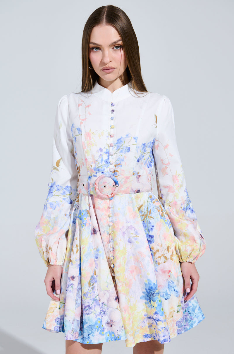 A woman elegantly poses in the EXPENSIVE TASTE FLORAL MINI DRESS, a luxurious skater-style mini adorned with pastel blue, pink, and yellow floral patterns. The dress features long sleeves and a matching belt that cinches her waist as she stands against a plain light background.