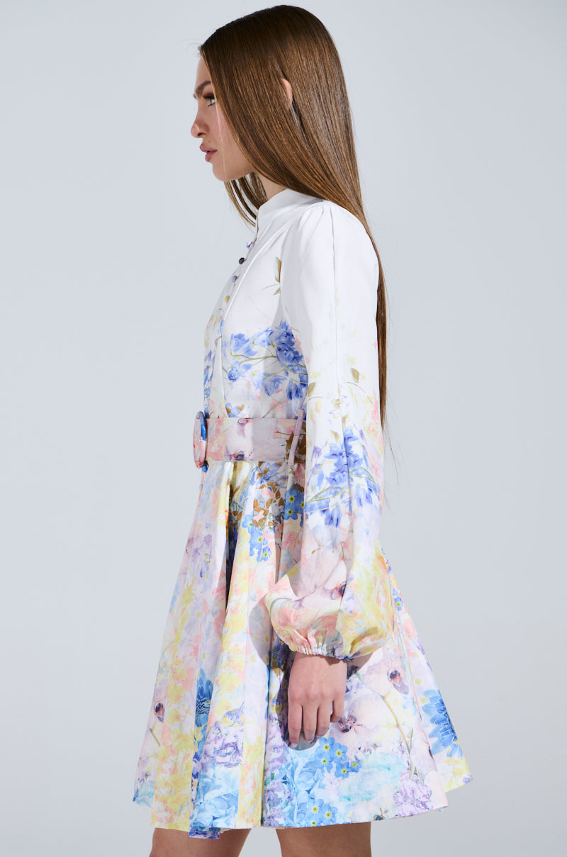 A person with long brown hair stands in profile, wearing the EXPENSIVE TASTE FLORAL MINI DRESS, a luxurious white floral mini dress featuring colorful patterns and long sleeves. The skater silhouette is cinched at the waist with a matching belt against a plain light gray background.
