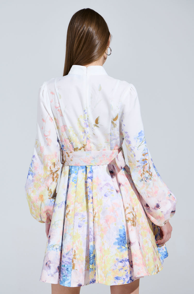 A person with long hair wears the EXPENSIVE TASTE FLORAL MINI DRESS, featuring puffy sleeves and pastel pink, blue, and yellow colors, set against a light gray backdrop.
