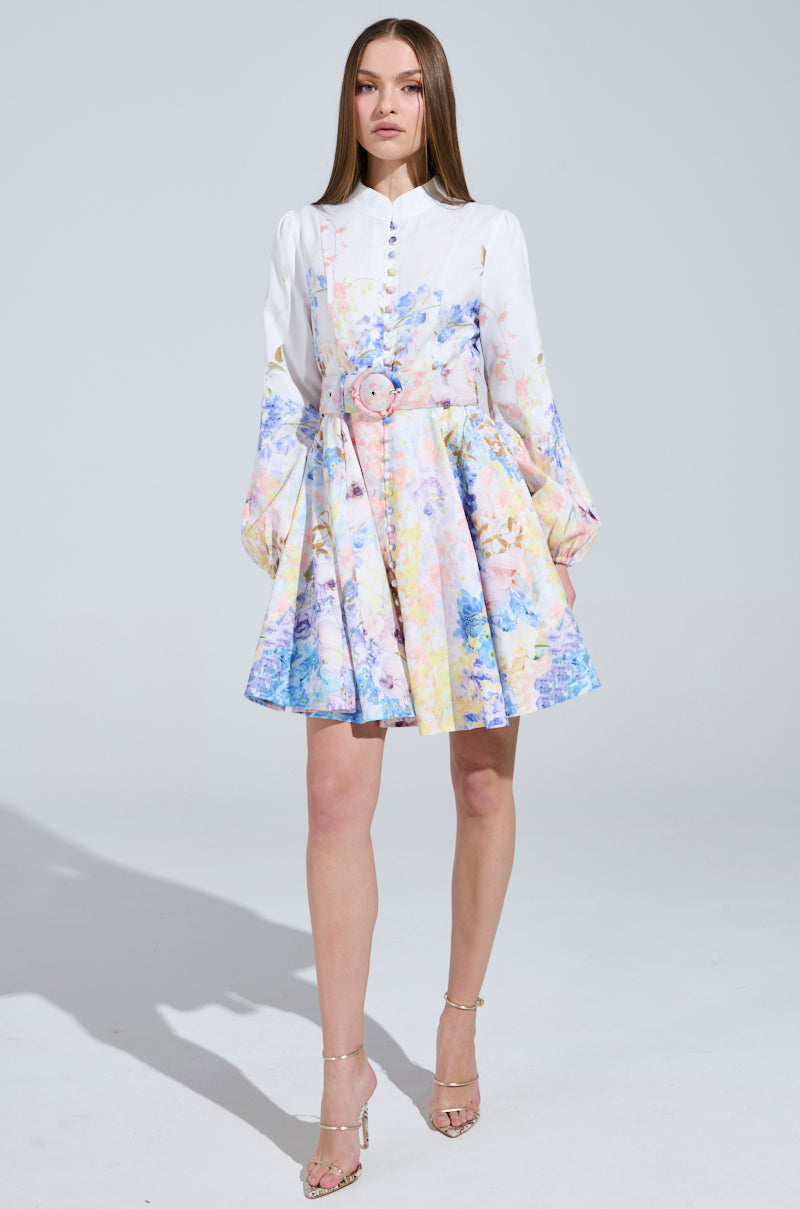 A woman stands elegantly in the EXPENSIVE TASTE FLORAL MINI DRESS, showcasing its floral pastel design and skater silhouette with long sleeves and a cinched waist with a belt. She pairs it with high-heeled sandals, confidently facing the camera against a plain backdrop.