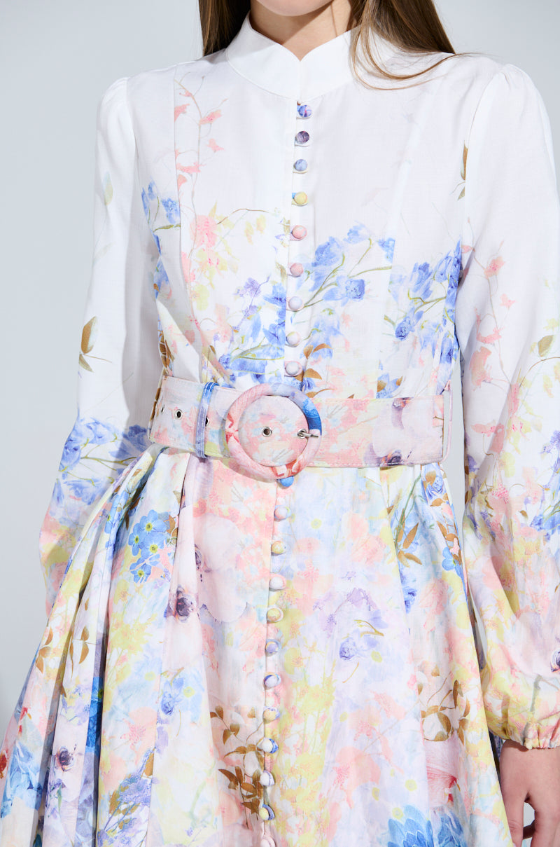 Dressed in the Expensive Taste Floral Mini Dress, featuring a long-sleeved floral pastel pattern and belt with a circular buckle, a person complements their long brown hair against a plain light gray background.